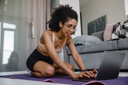 Photo for Black young woman wearing eyeglasses using laptop during workout at home - Royalty Free Image