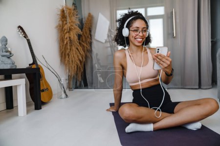 Foto de Mujer joven afroamericana escuchando música con auriculares mientras practica yoga en casa - Imagen libre de derechos