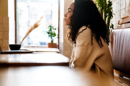 Téléchargez les photos : Jeune femme noire travaillant avec un ordinateur portable tout en étant assis dans un café à l'intérieur - en image libre de droit