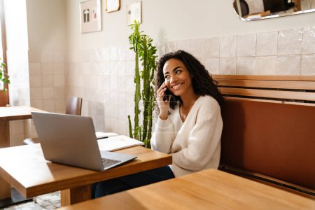 Téléchargez les photos : Jeune femme noire parlant sur un téléphone portable tout en travaillant avec un ordinateur portable dans un café à l'intérieur - en image libre de droit