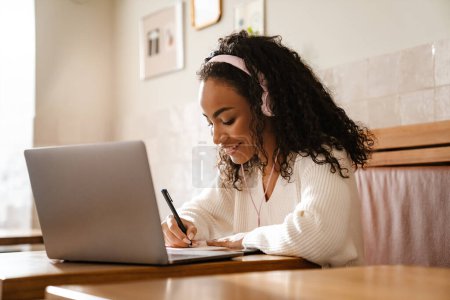 Téléchargez les photos : Jeune femme noire note tout en travaillant avec un ordinateur portable dans un café à l'intérieur - en image libre de droit