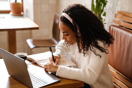 Téléchargez les photos : Jeune femme noire note tout en travaillant avec un ordinateur portable dans un café à l'intérieur - en image libre de droit