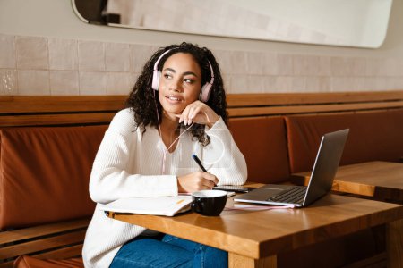 Foto de Joven mujer negra escribiendo notas mientras trabaja con el ordenador portátil en el café en el interior - Imagen libre de derechos