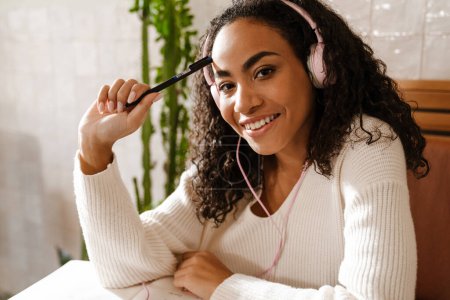 Téléchargez les photos : Jeune femme noire écouter de la musique tout en écrivant des notes dans le café à l'intérieur - en image libre de droit