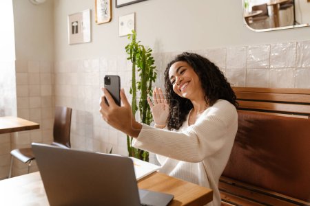Téléchargez les photos : Jeune femme noire gestuelle et prendre selfie photo sur téléphone portable dans le café à l'intérieur - en image libre de droit