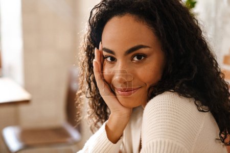 Foto de Joven mujer negra sonriendo y mirando a la cámara mientras está sentado en el café en el interior - Imagen libre de derechos