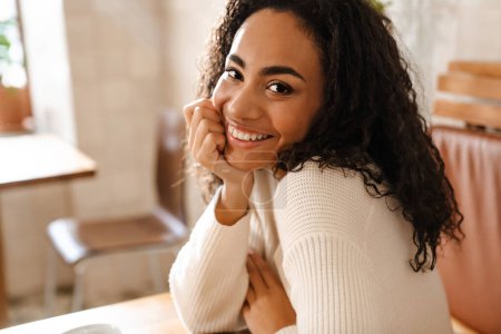 Téléchargez les photos : Jeune femme noire souriant et regardant la caméra tout en étant assis dans un café à l'intérieur - en image libre de droit