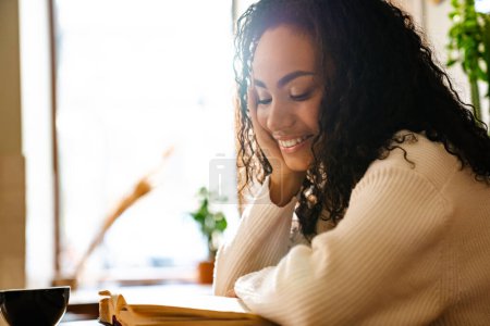 Foto de Joven mujer negra sonriendo y leyendo libro en la cafetería en el interior - Imagen libre de derechos