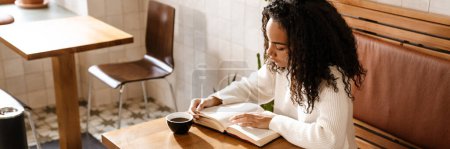 Photo for Young black woman drinking coffee and reading book in cafe indoors - Royalty Free Image