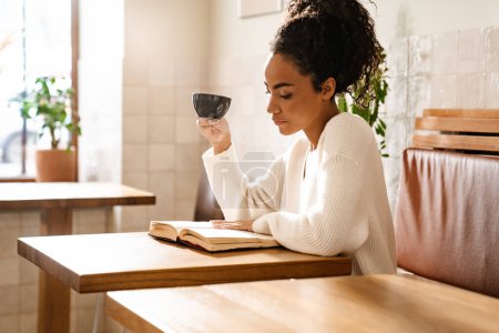 Foto de Joven mujer negra bebiendo café y leyendo libro en la cafetería interior - Imagen libre de derechos