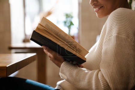 Foto de Joven mujer negra sonriendo y leyendo libro en la cafetería en el interior - Imagen libre de derechos