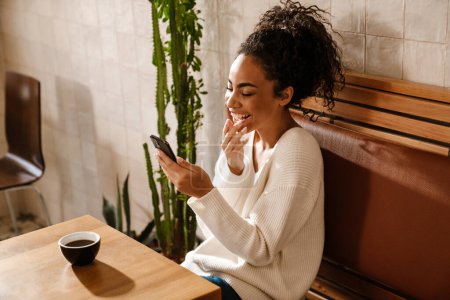 Foto de Joven mujer negra usando el teléfono móvil mientras bebe café en el café en el interior - Imagen libre de derechos
