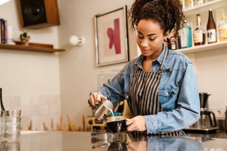 Téléchargez les photos : Jeune barista noire portant tablier faisant du café tout en travaillant dans un café à l'intérieur - en image libre de droit