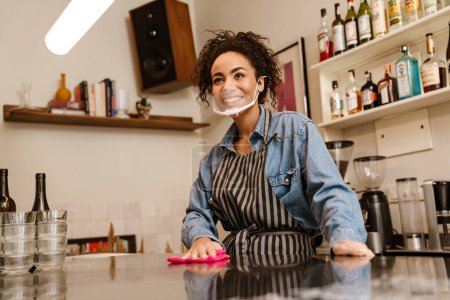 Foto de Mujer barista negra con máscara facial sonriendo mientras trabaja en un café en el interior - Imagen libre de derechos