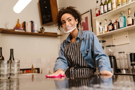 Téléchargez les photos : Femme barista noire portant un masque facial souriant tout en travaillant dans un café à l'intérieur - en image libre de droit