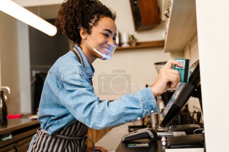 Foto de Negro barista mujer usando mascarilla de trabajo con caja registradora en el café en interiores - Imagen libre de derechos
