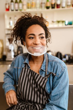 Foto de Joven camarera negra con delantal y mascarilla sonriendo mientras trabaja en la cafetería en interiores - Imagen libre de derechos
