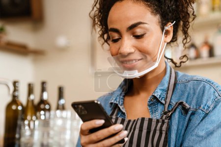 Téléchargez les photos : Jeune serveuse noire en masque facial utilisant un téléphone portable tout en travaillant au café à l'intérieur - en image libre de droit