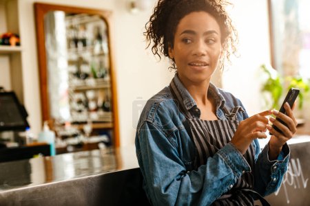 Téléchargez les photos : Jeune serveuse noire utilisant un téléphone portable tout en travaillant au café à l'intérieur - en image libre de droit