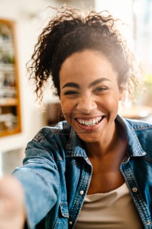 Téléchargez les photos : Jeune serveuse noire souriante et prenant des photos selfie tout en travaillant au café à l'intérieur - en image libre de droit
