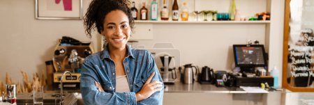 Téléchargez les photos : Jeune serveuse noire portant une chemise souriante tout en travaillant au café à l'intérieur - en image libre de droit