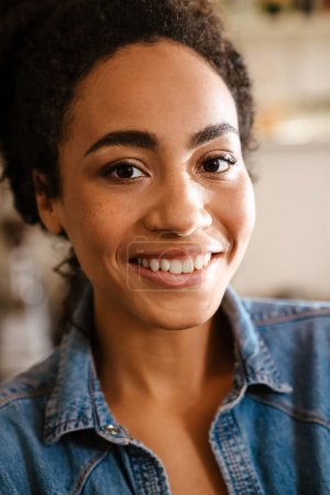 Téléchargez les photos : Jeune femme noire portant une chemise en denim souriant et regardant la caméra à l'intérieur - en image libre de droit