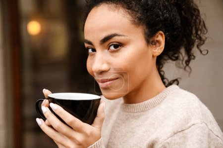 Téléchargez les photos : Jeune femme noire souriant et buvant du café dans un café à l'extérieur - en image libre de droit