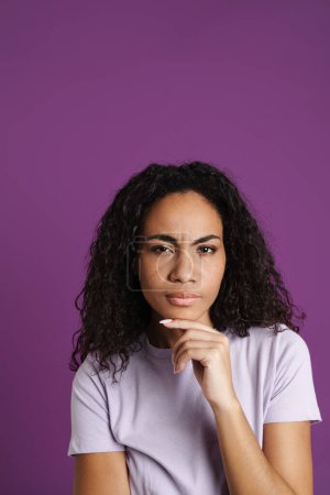 Foto de Joven mujer negra con el pelo ondulado frunciendo el ceño y mirando a la cámara aislada sobre fondo púrpura - Imagen libre de derechos