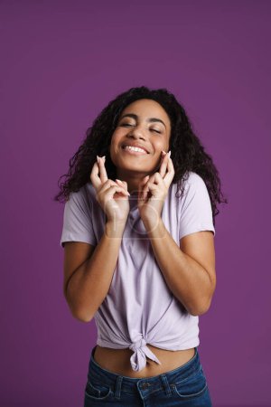 Foto de Joven mujer negra sosteniendo los dedos cruzados para la buena suerte aislado sobre fondo púrpura - Imagen libre de derechos