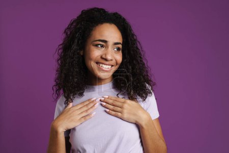 Foto de Joven mujer negra riendo mientras toma las manos sobre su pecho aislado sobre fondo púrpura - Imagen libre de derechos