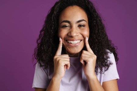 Foto de Joven mujer negra con el pelo ondulado señalando con los dedos su sonrisa aislada sobre el fondo púrpura - Imagen libre de derechos