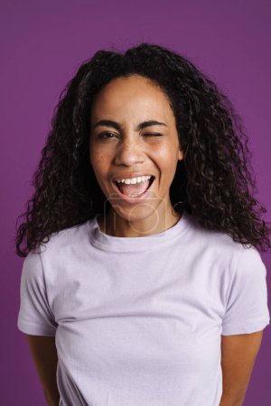 Téléchargez les photos : Jeune femme noire avec des cheveux ondulés clin d'oeil et souriant à la caméra isolé sur fond violet - en image libre de droit