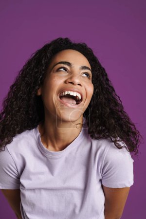 Téléchargez les photos : Jeune femme noire aux cheveux ondulés riant à la caméra isolée sur fond violet - en image libre de droit