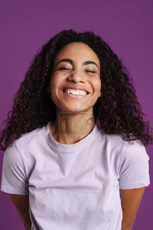 Téléchargez les photos : Jeune femme noire aux cheveux ondulés souriant à la caméra isolée sur fond violet - en image libre de droit