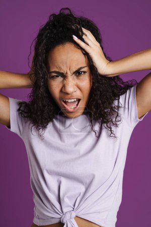 Photo for Furious black woman with wavy hair screaming while holding her head isolated over purple background - Royalty Free Image