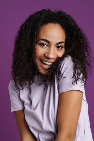 Téléchargez les photos : Jeune femme noire aux cheveux ondulés riant et regardant la caméra isolée sur fond violet - en image libre de droit