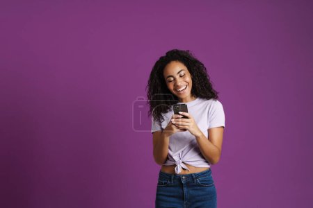Foto de Joven mujer negra usando teléfono móvil y riendo aislado sobre fondo púrpura - Imagen libre de derechos