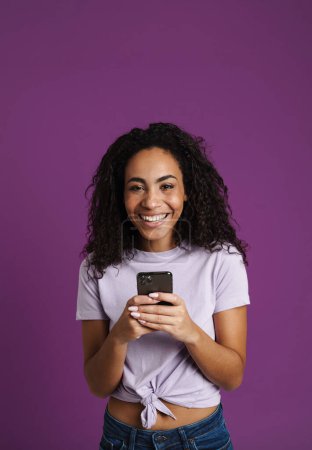 Téléchargez les photos : Jeune femme noire utilisant le téléphone mobile et riant isolé sur fond violet - en image libre de droit