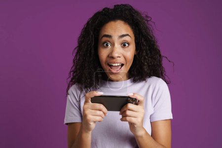 Téléchargez les photos : Surpris femme noire jouer jeu en ligne sur téléphone mobile isolé sur fond violet - en image libre de droit