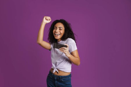 Téléchargez les photos : Jeune femme noire gestuelle tout en jouant jeu en ligne sur téléphone cellulaire isolé sur fond violet - en image libre de droit