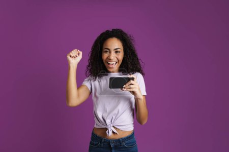 Téléchargez les photos : Jeune femme noire gestuelle tout en jouant jeu en ligne sur téléphone cellulaire isolé sur fond violet - en image libre de droit