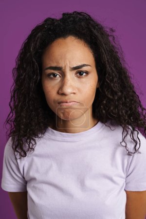 Téléchargez les photos : Jeune femme noire avec les cheveux ondulés fronçant les sourcils et regardant la caméra isolée sur fond violet - en image libre de droit
