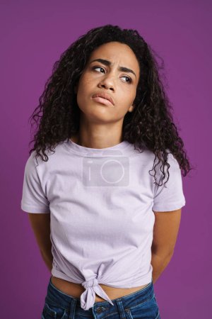 Foto de Joven mujer negra con el pelo ondulado frunciendo el ceño y mirando a un lado aislado sobre fondo púrpura - Imagen libre de derechos