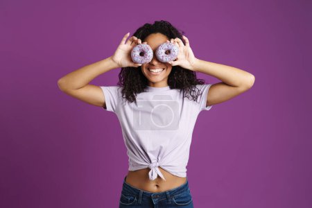 Foto de Joven mujer negra usando camiseta burlándose con rosquillas aisladas sobre fondo púrpura - Imagen libre de derechos