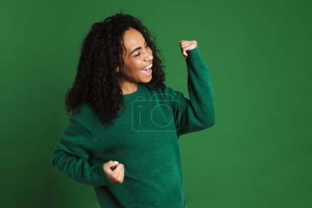 Téléchargez les photos : Jeune femme noire riant tout en faisant geste gagnant isolé sur fond vert - en image libre de droit