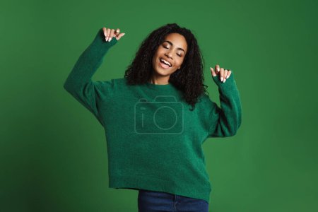 Foto de Joven mujer negra con el pelo ondulado sonriendo y bailando aislado sobre fondo verde - Imagen libre de derechos