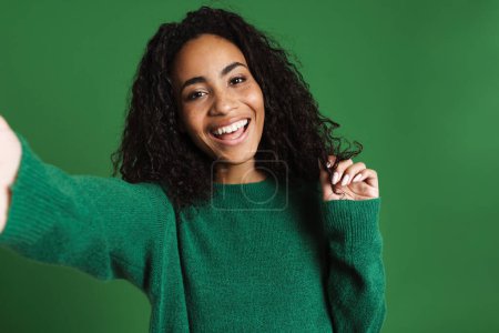 Téléchargez les photos : Jeune femme noire portant pull riant tout en prenant selfie photo isolé sur fond vert - en image libre de droit
