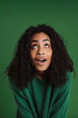 Foto de Joven mujer negra con el pelo ondulado expresando sorpresa ante la cámara aislada sobre fondo verde - Imagen libre de derechos