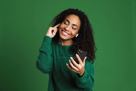 Foto de Joven mujer negra usando el teléfono móvil mientras escucha música aislada sobre fondo verde - Imagen libre de derechos