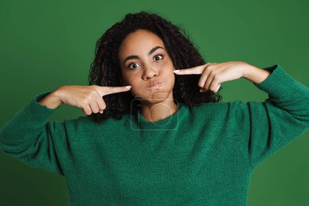 Foto de Joven mujer negra haciendo muecas mientras señala con los dedos sus mejillas aisladas sobre un fondo verde - Imagen libre de derechos